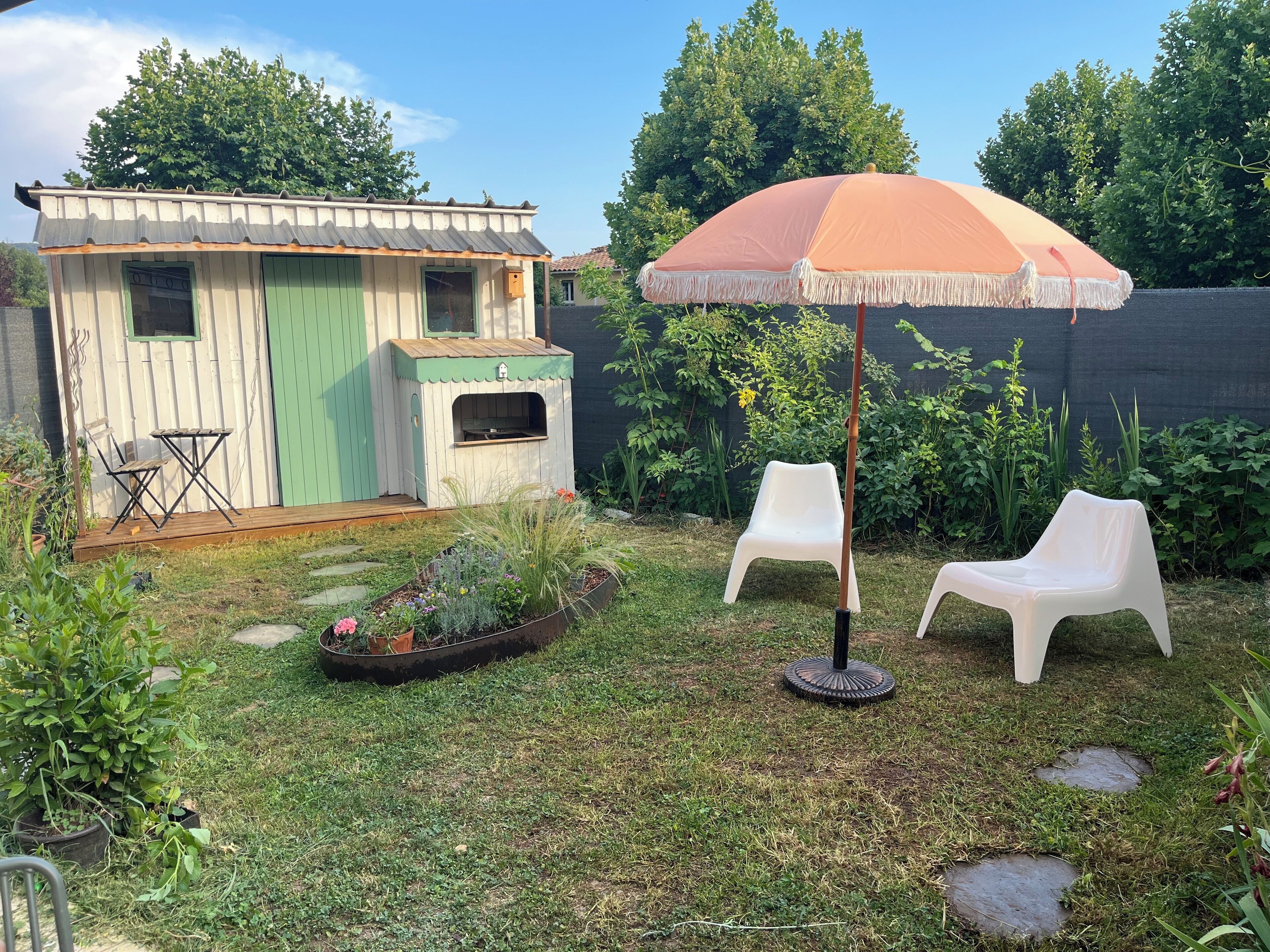 A backyard with a wooden shed, a barbecue grill, two white plastic chairs, and an umbrella.
