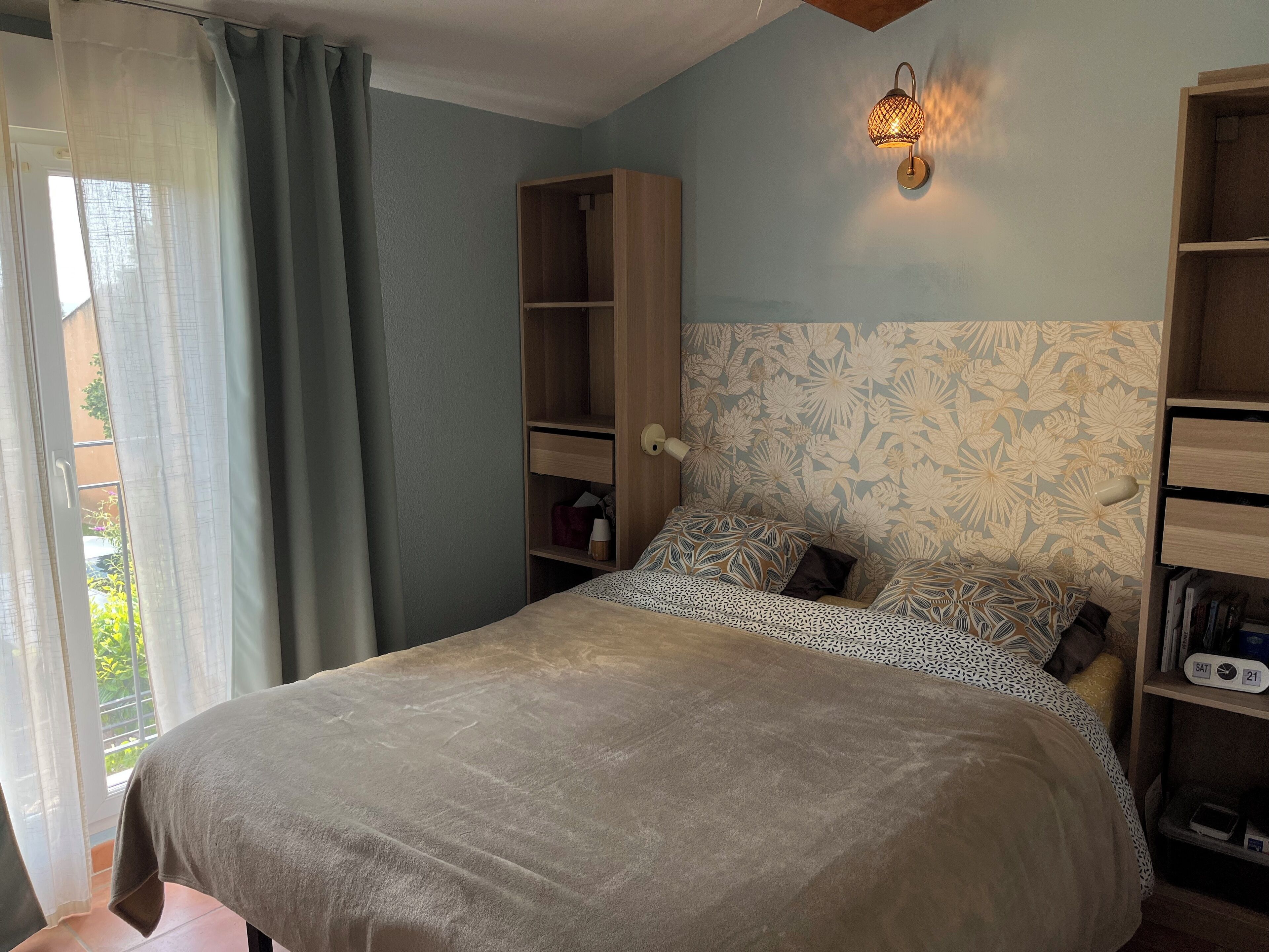 A bedroom with a bed, a wardrobe, a nightstand, a wall-mounted lamp, and a window with curtains.