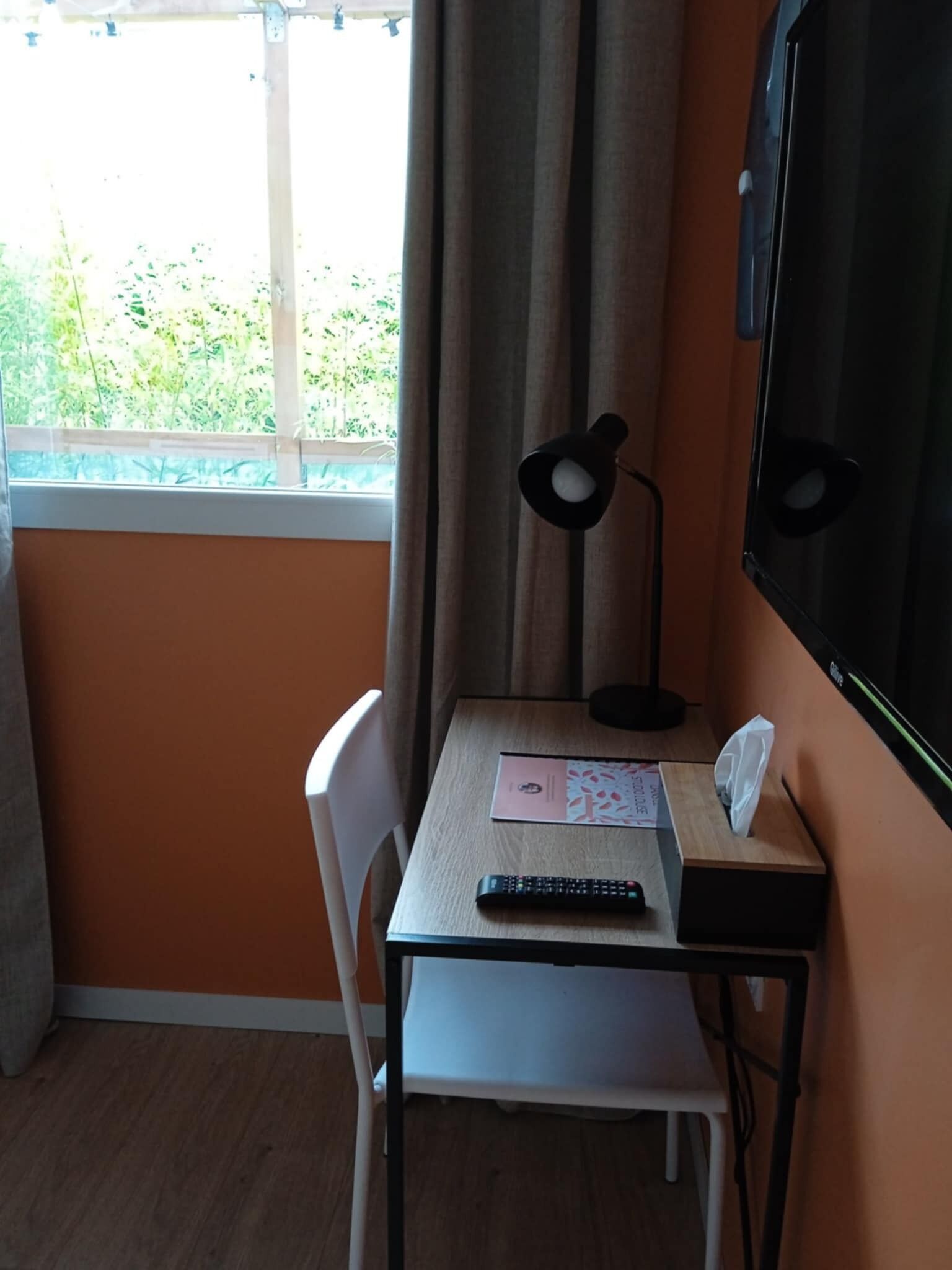 A small room with a single table, a chair, a desk lamp, a remote control, and a television.