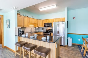 A modern kitchen with wooden cabinets, a stainless steel refrigerator, a microwave, and a bar area with stools. - Pet-friendly, centrally-located oceanside condo w/ community pool! (Kill Devil Hills)