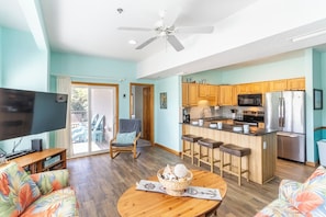 A living room with a TV, sofa, and coffee table, a kitchen with wooden cabinets and a refrigerator, and a balcony with a chair.