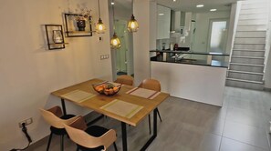 Un comedor moderno con una mesa de madera, sillas y un frutero. Se ve una cocina al fondo, separada por un panel de vidrio.