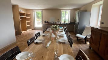 A dining room with a long wooden table set for six, surrounded by chairs, with a kitchen and a window in the background.
