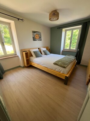 A bedroom with a wooden bed, two pillows, a bedside table, a hanging light fixture, and a window with curtains.