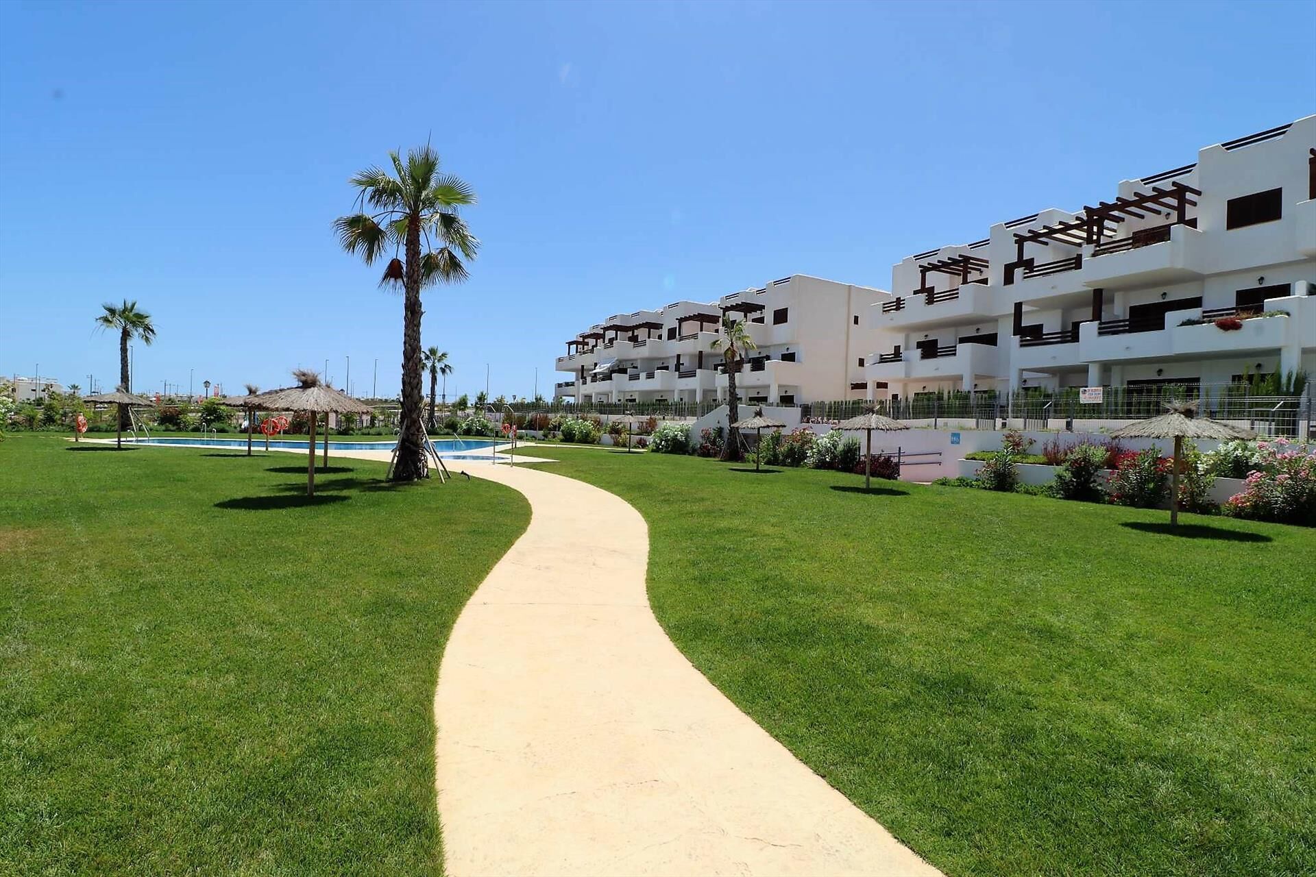 Un sendero conduce a un edificio moderno con varios balcones y un área de piscina con palmeras.