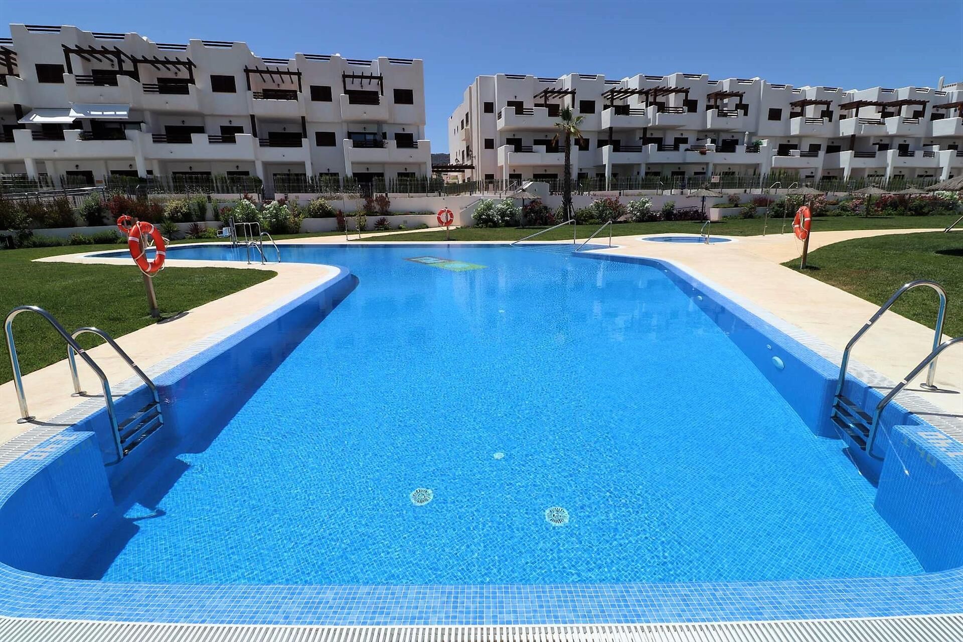 A rectangular swimming pool with a blue tiled floor, surrounded by a low concrete deck. There are two lifebuoys and metal handrails on the deck. In the background, there are multi-story buildings with balconies and a clear sky.