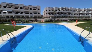 A rectangular swimming pool with a blue tiled floor, surrounded by a low concrete deck. There are two lifebuoys and metal handrails on the deck. In the background, there are multi-story buildings with balconies and a clear sky. - Casa Soleada - Three Bedroom Apartment, Sleeps 6 (Pulpí)