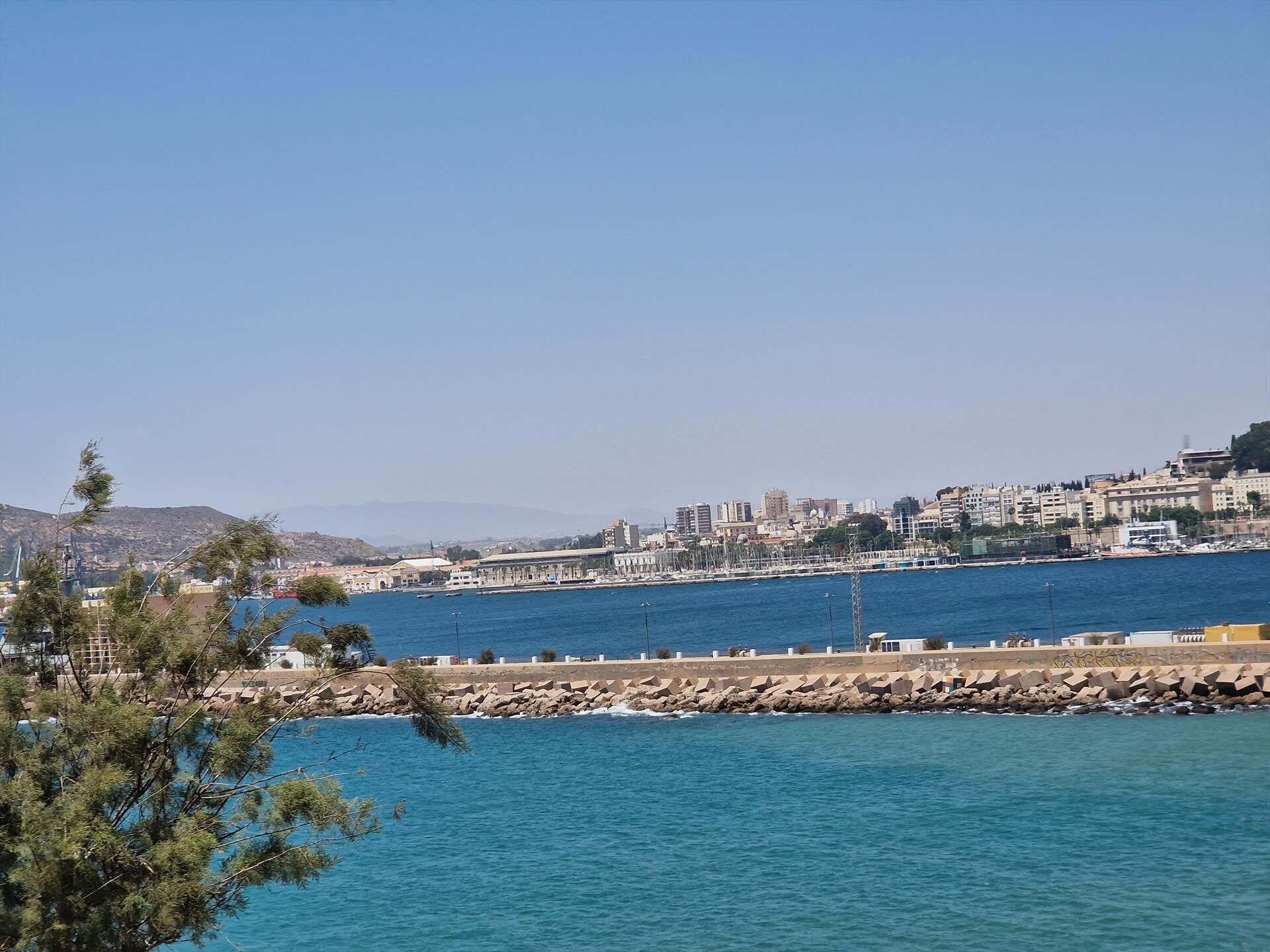 Un paisaje urbano costero con un espigón rocoso, mar azul cristalino y una combinación de edificios residenciales y comerciales.