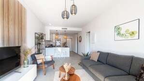 Un salon moderne avec un canapé gris, une table basse et un téléviseur posé sur un meuble blanc. On aperçoit une cuisine à l’arrière-plan, avec un coin repas.