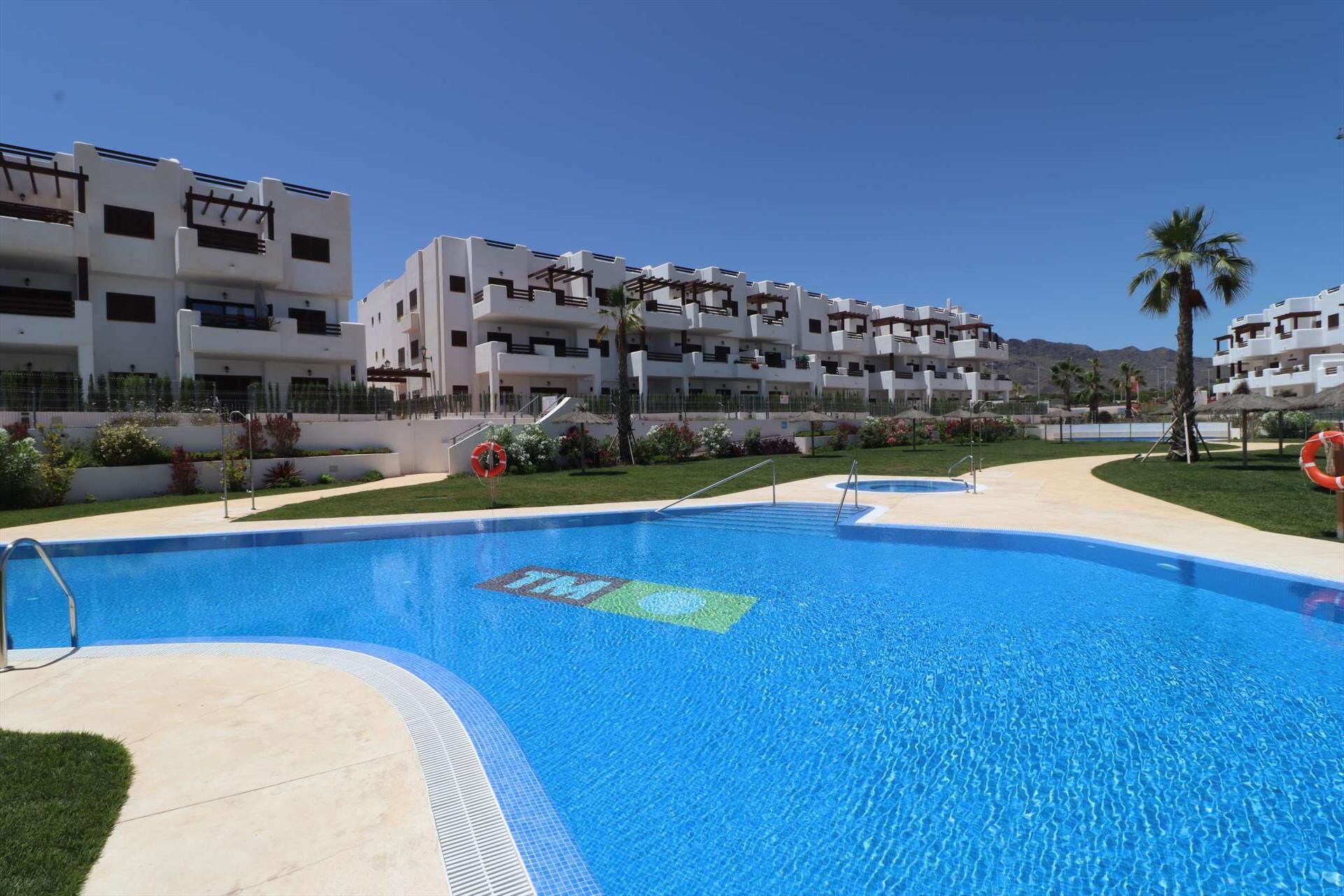 Una piscina con un flotador salvavidas, rodeada de un césped cuidado y un edificio con balcones.