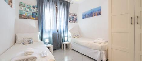 A hotel room with two beds, a wardrobe, a small table, and a window with curtains.