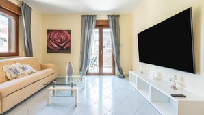 A living room with a sofa, a glass coffee table, a TV on the wall, and a balcony door. - Holiday Home 'P012' with Balcony, Wi-Fi and Air Conditioning (Cagliari)