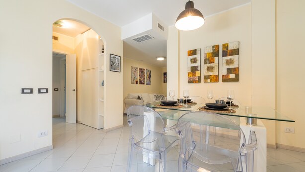 A modern kitchen with a glass dining table and chairs, a hanging light fixture, and artwork on the walls. - Holiday Home 'P012' with Balcony, Wi-Fi and Air Conditioning (Cagliari)