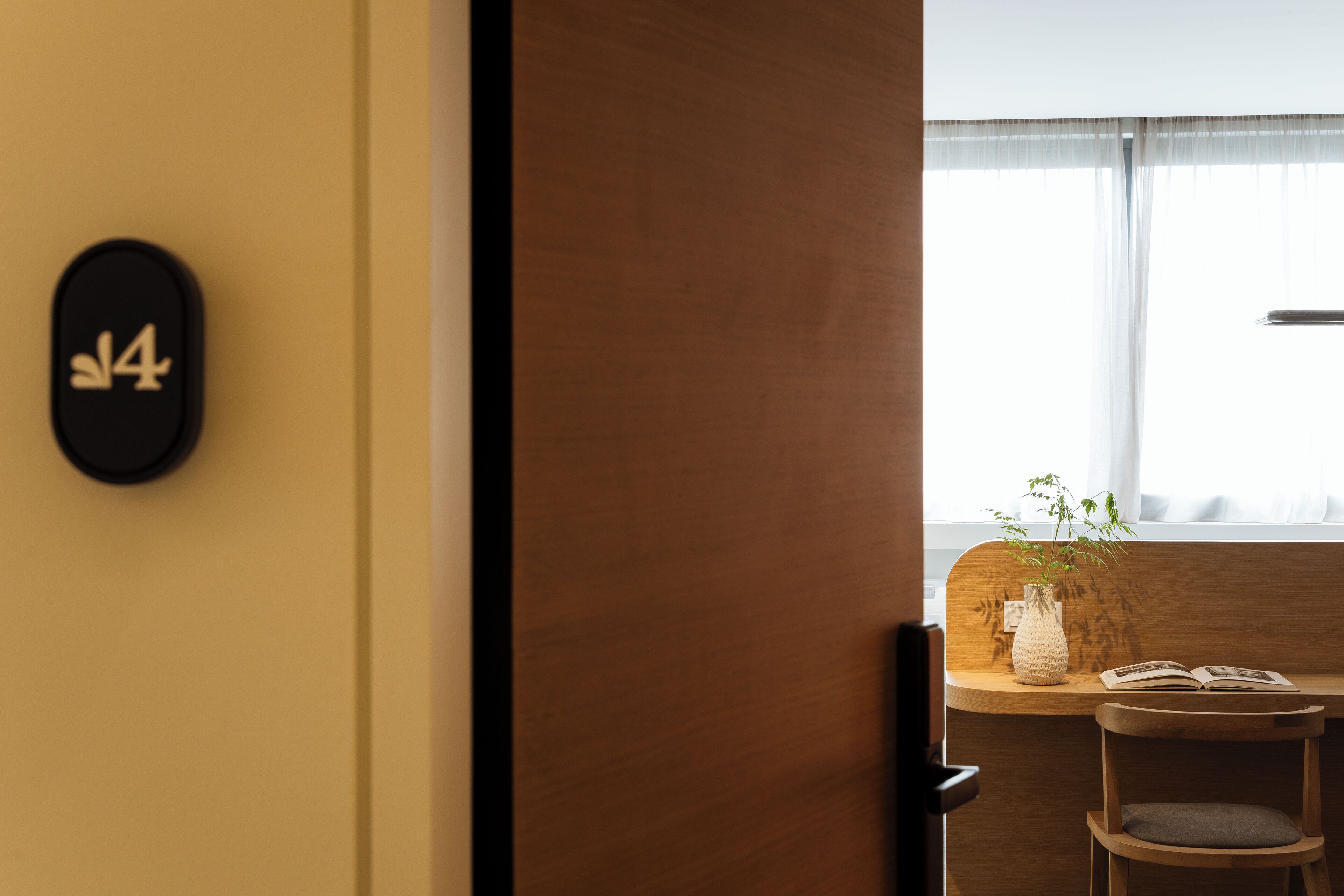 Una habitación con escritorio de madera, una silla, planta en maceta y ventana con cortinas traslúcidas.