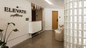 Ein moderner Empfangsbereich mit einem Schild mit der Aufschrift "ELEVATE Athens", einem weißen Empfangstresen, einer Holztür und einer Glastrennwand.