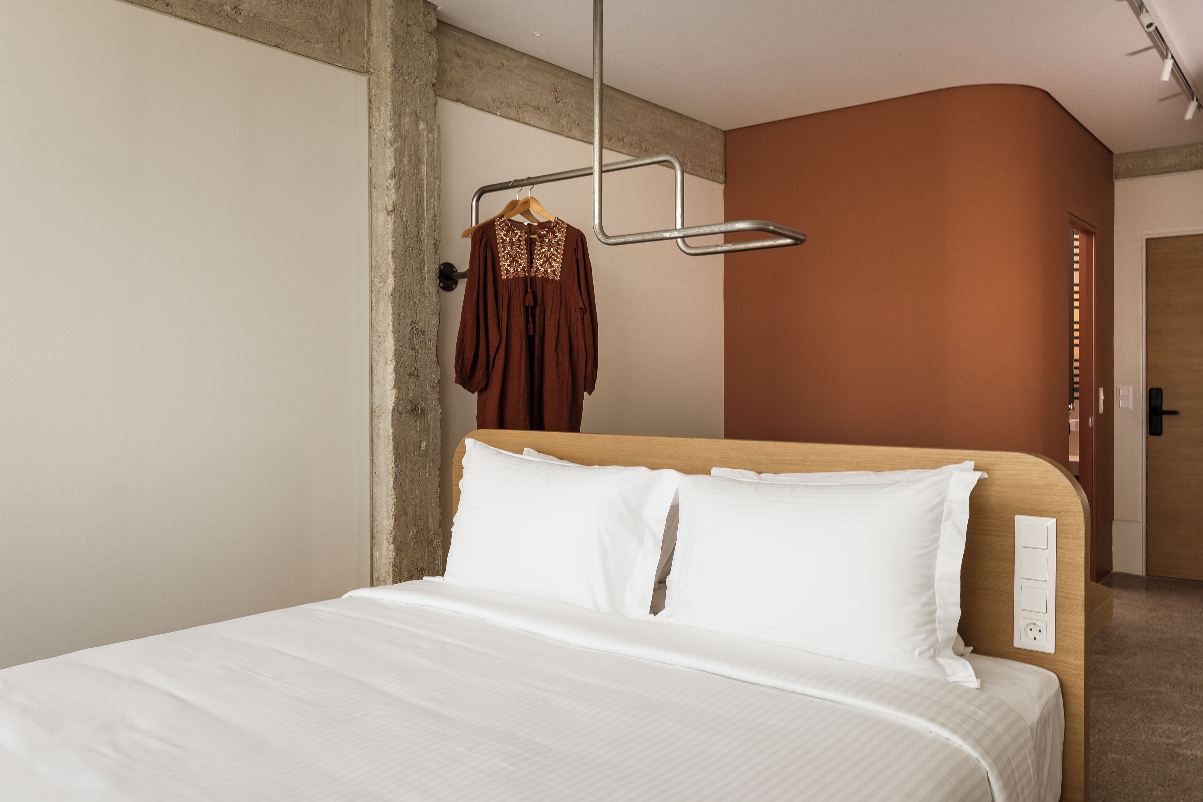 A modern hotel room with a wooden headboard, white bedding, and a hanging garment rack.