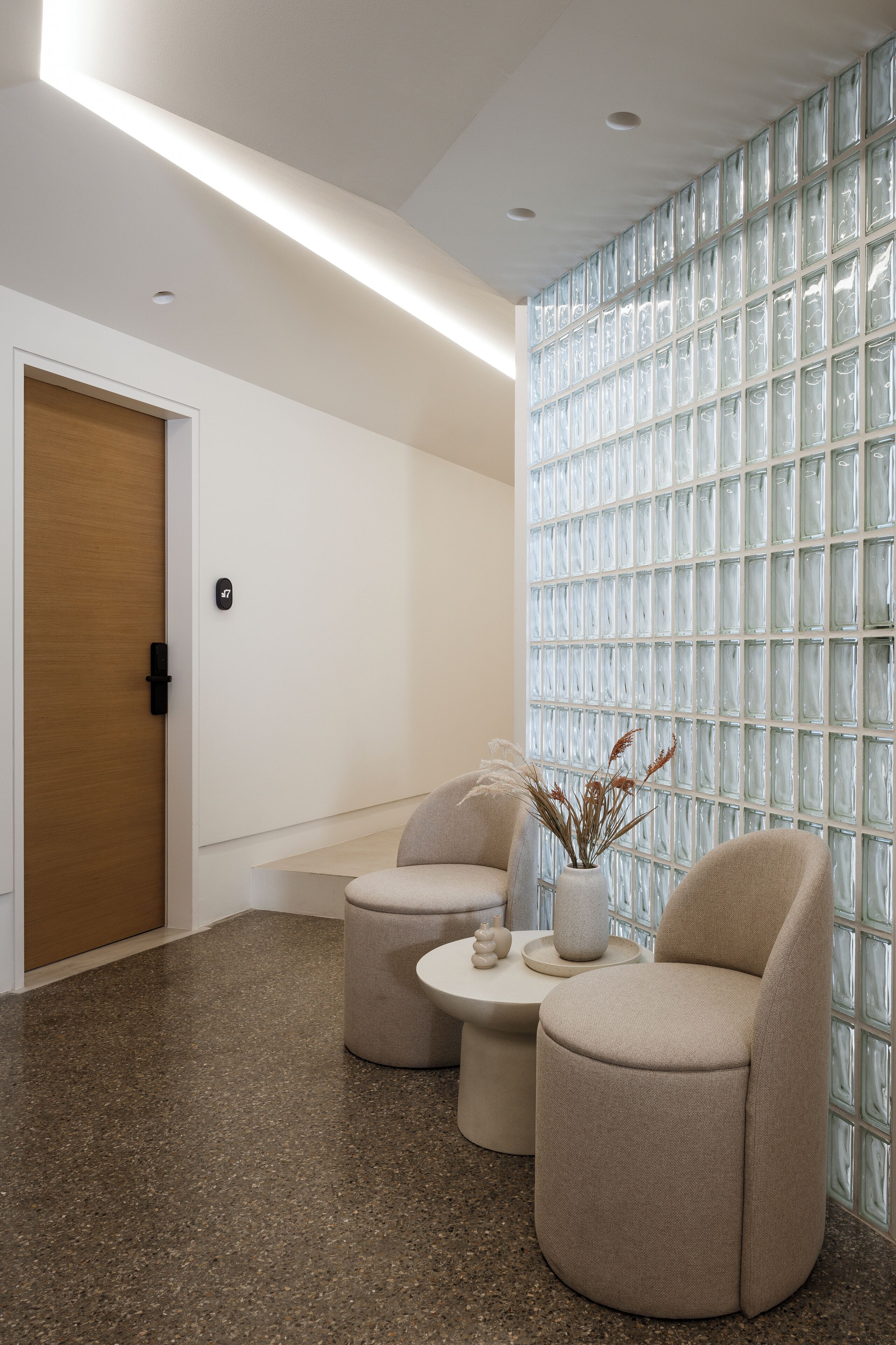 A modern waiting area with a wooden door, a glass partition wall, and two beige armchairs with a small table and vase between them.