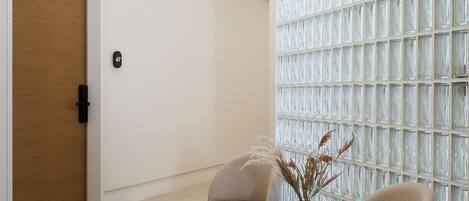 A modern waiting area with a wooden door, a glass partition wall, and two beige armchairs with a small table and vase between them.