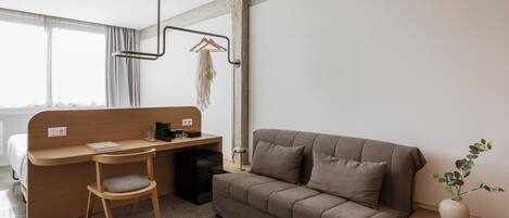 A modern hotel room with a wooden desk, a sofa, a hanging plant, and a view through large windows.
