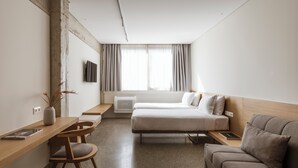 Ein modernes Hotelzimmer mit Schreibtisch aus Holz, zwei Einzelbetten, einer Couch und einem großen Fenster mit Vorhängen.