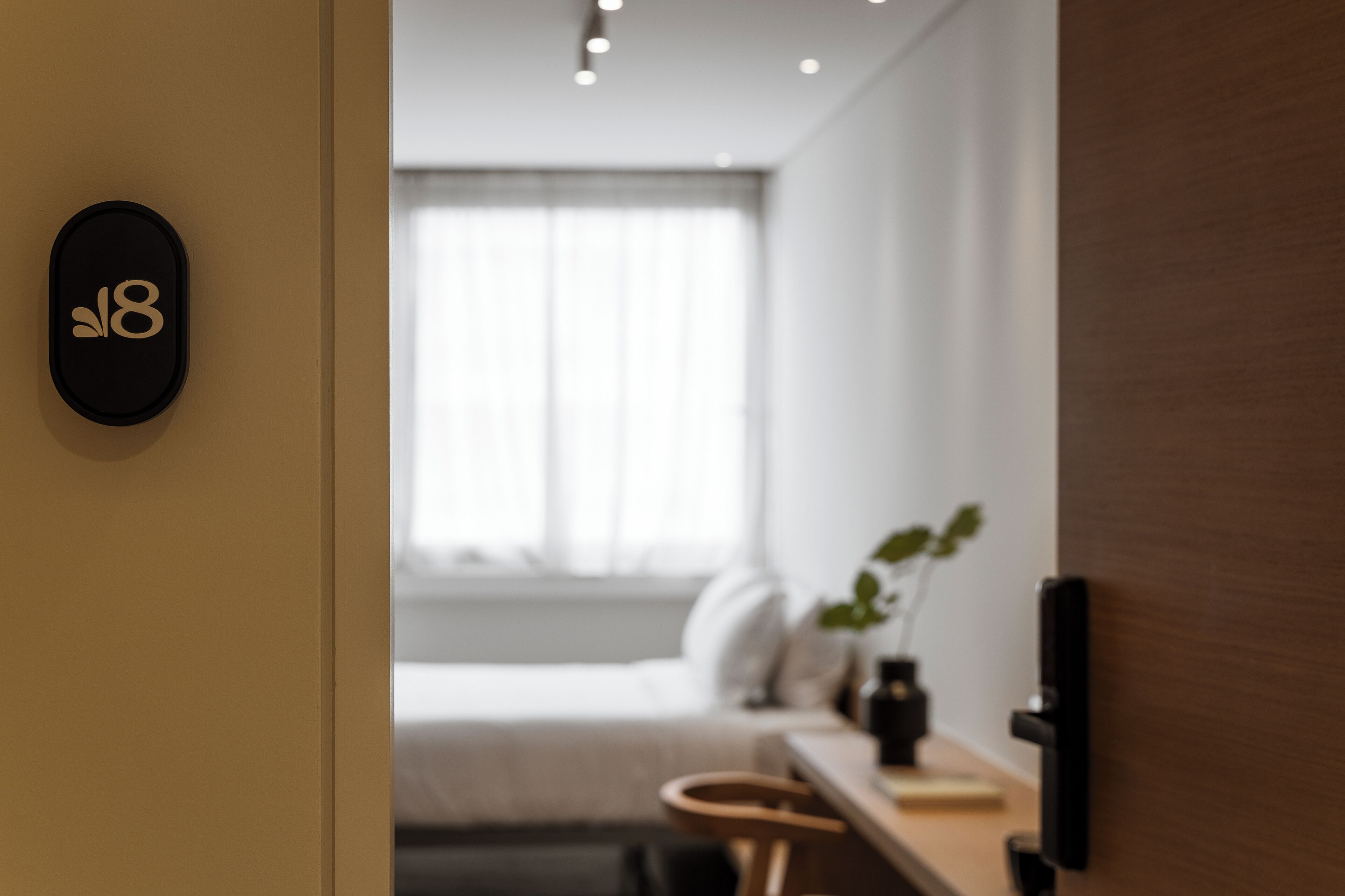 A modern hotel room with a minimalist design, featuring a bed, a desk, and a potted plant.