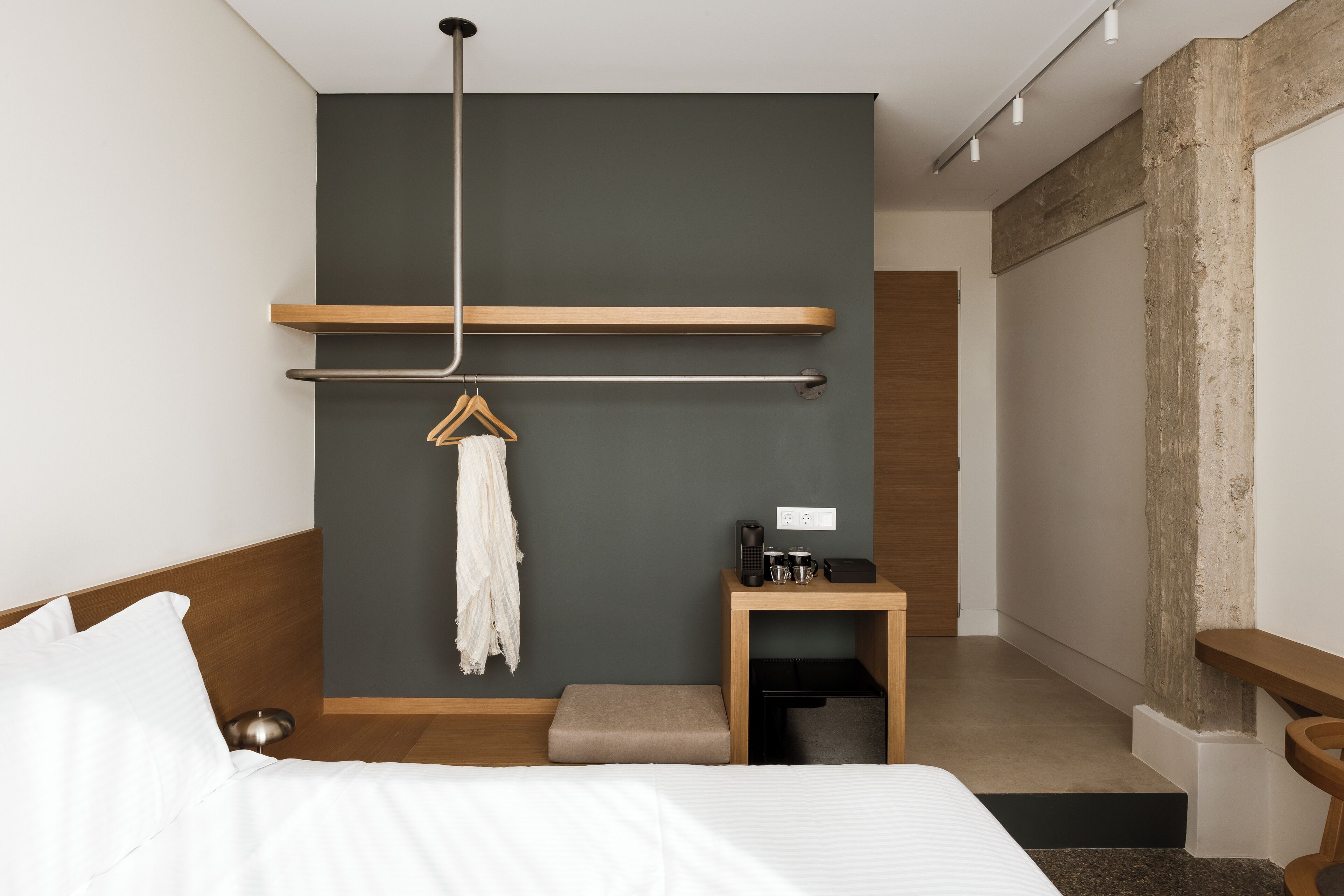 A modern hotel room with a bed, a small bedside table, a hanging towel, and a wooden bench.