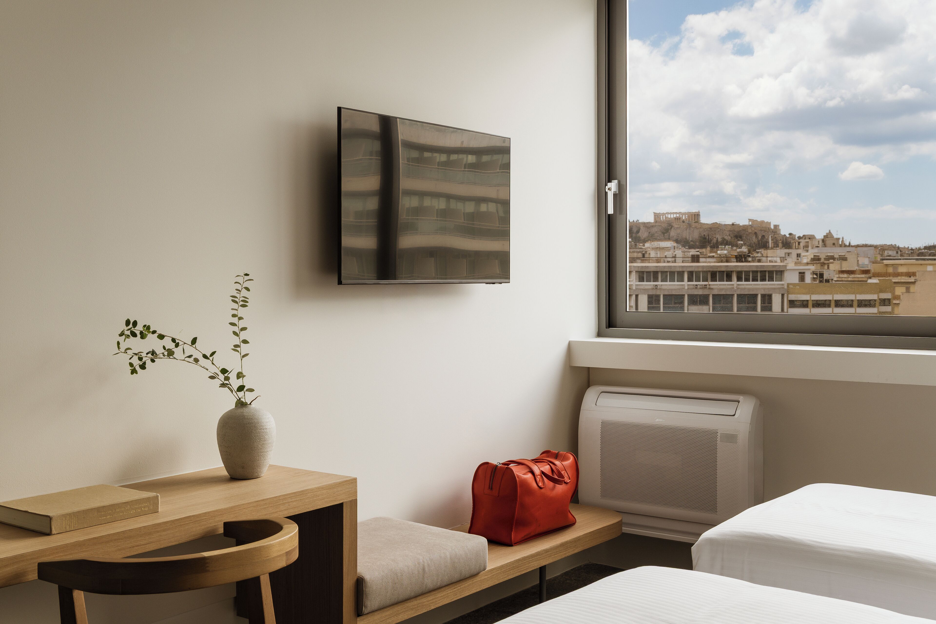 Hotelkamer met een bed, bureau met een boek, vaas met plant, rode tas, televisie aan de muur en raam met uitzicht op de stad.