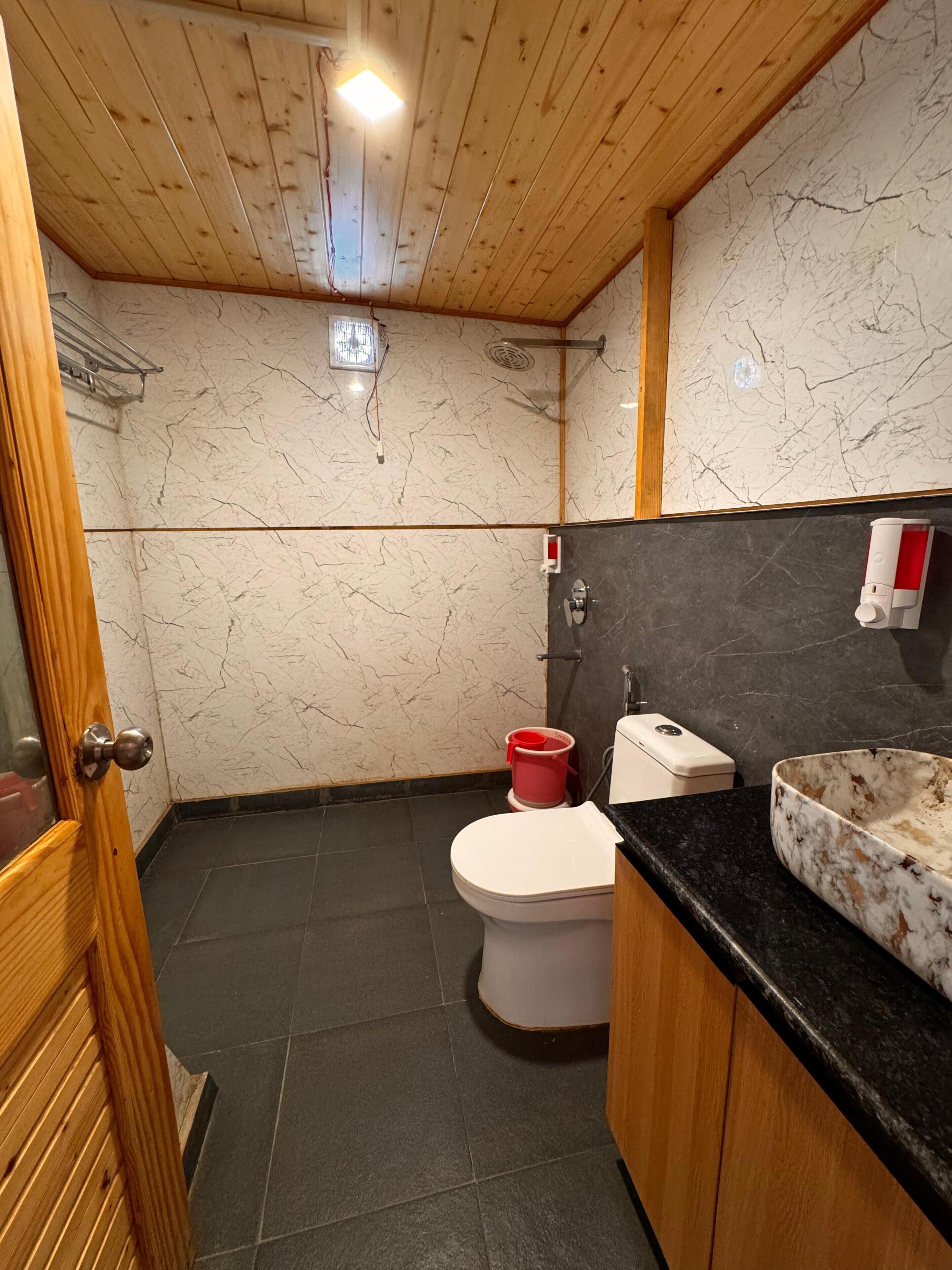 Un baño con techo de madera, paredes de mármol, un inodoro, un lavabo con lavamanos de mármol y un cubo rojo.
