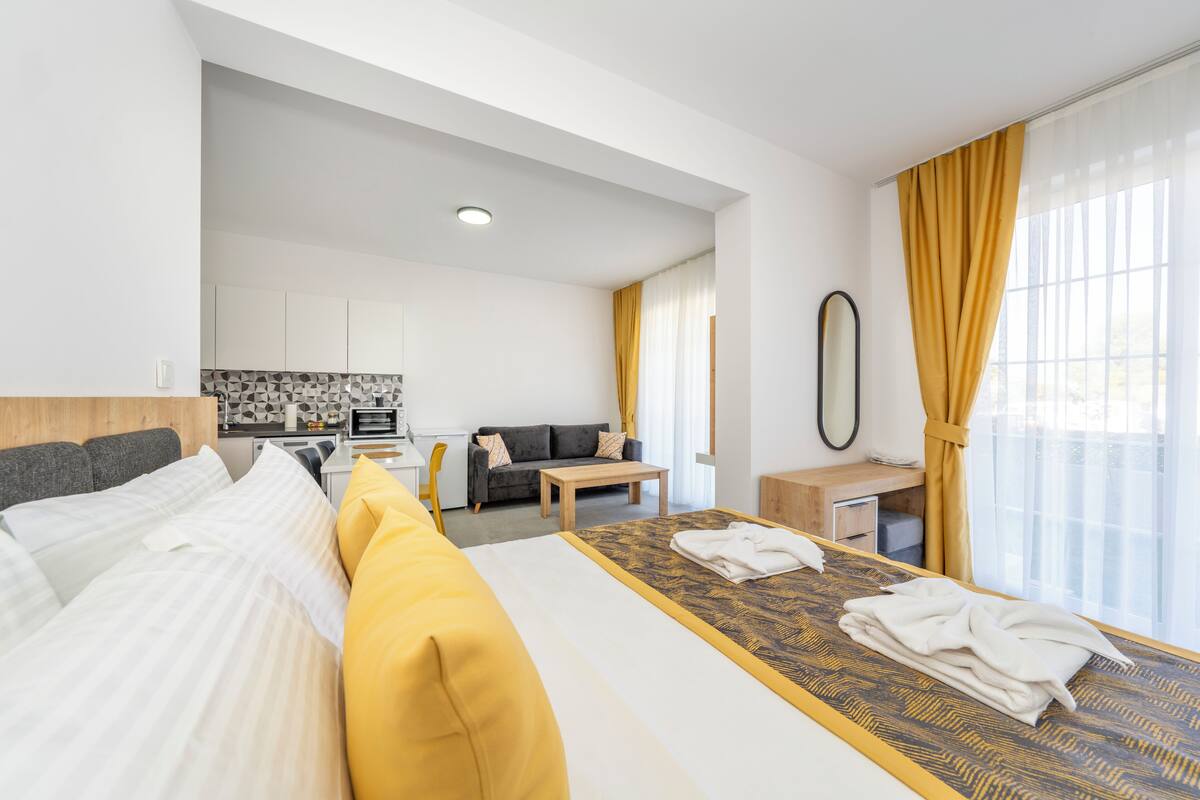 A modern hotel room with a large bed, a small desk, a sofa, and a kitchenette in the background.