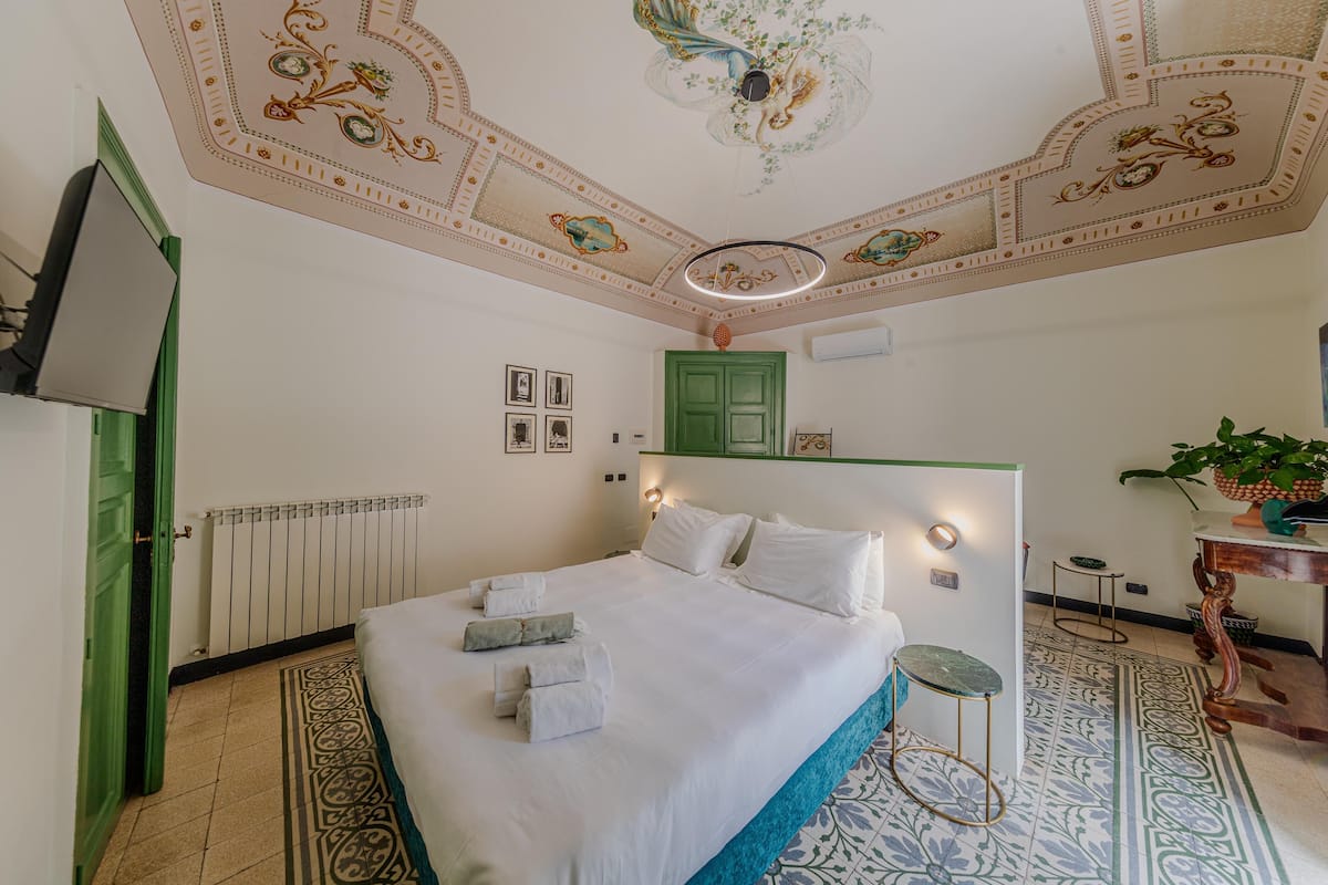 Habitación de hotel con una cama grande, una puerta verde, un radiador, un techo con motivos florales y una pequeña mesa con una planta.