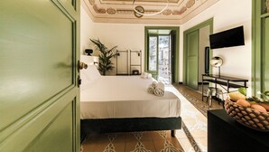 A hotel room with a bed, a desk, a television, and a view of the city through a green door. - Palazzo Villelmi (Cefalù)