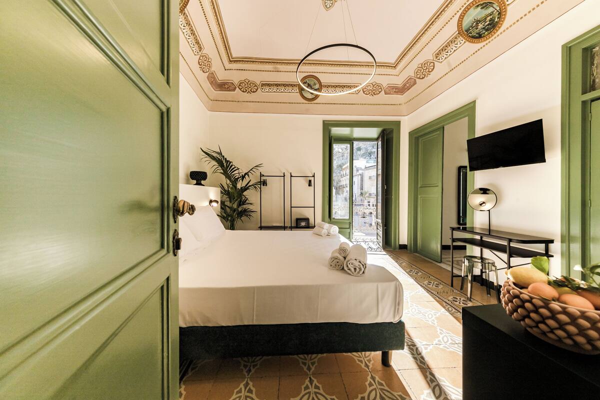Habitación de hotel con cama, escritorio, televisor y vista a la ciudad a través de una puerta verde.
