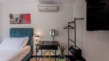 Una habitación de hotel moderna con una cama con cabecera color turquesa, un escritorio pequeño con espejo, una planta en maceta y un televisor montado en la pared.