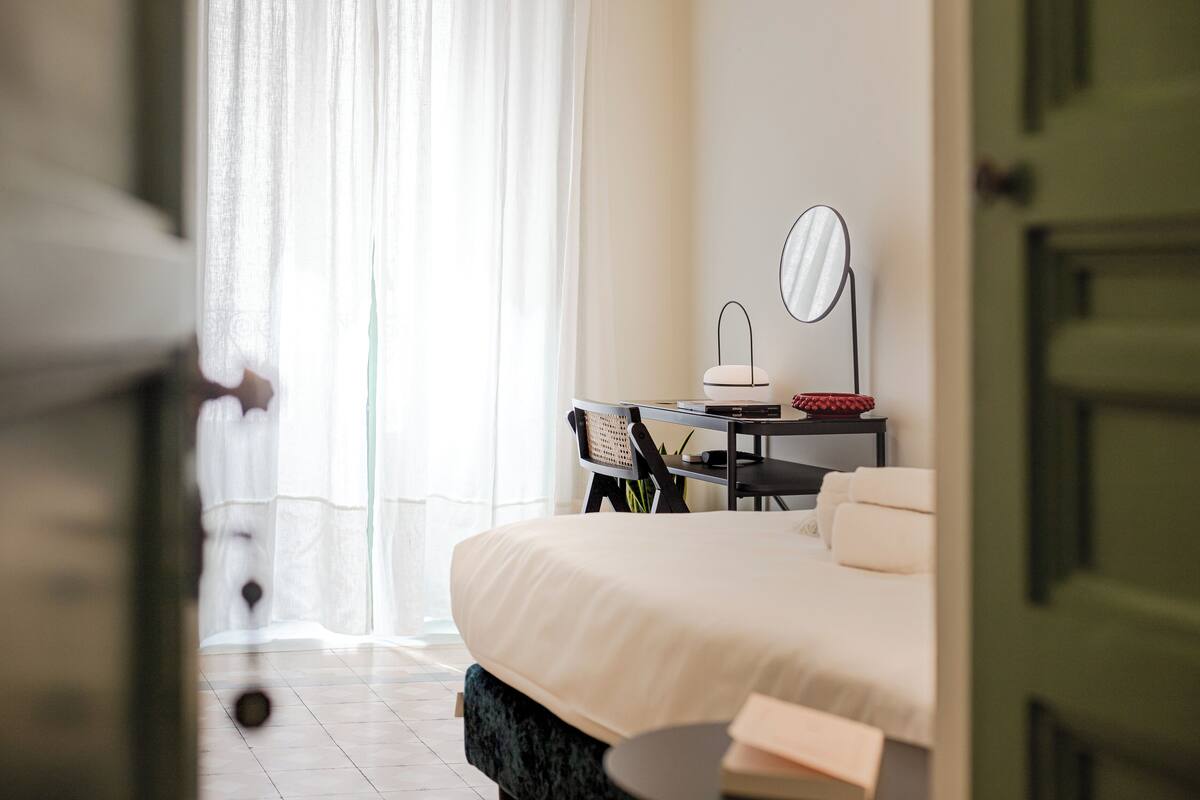 Un dormitorio moderno con una cama grande, una mesita de noche, una silla, un espejo y una ventana con cortinas de gasa.