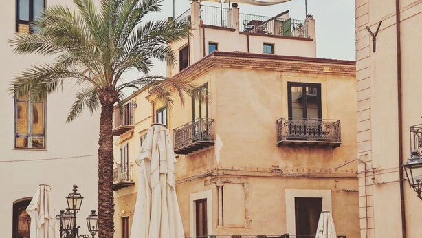 An outdoor cafe with tables and chairs, surrounded by buildings with balconies and palm trees. - Palazzo Villelmi (Cefalù)