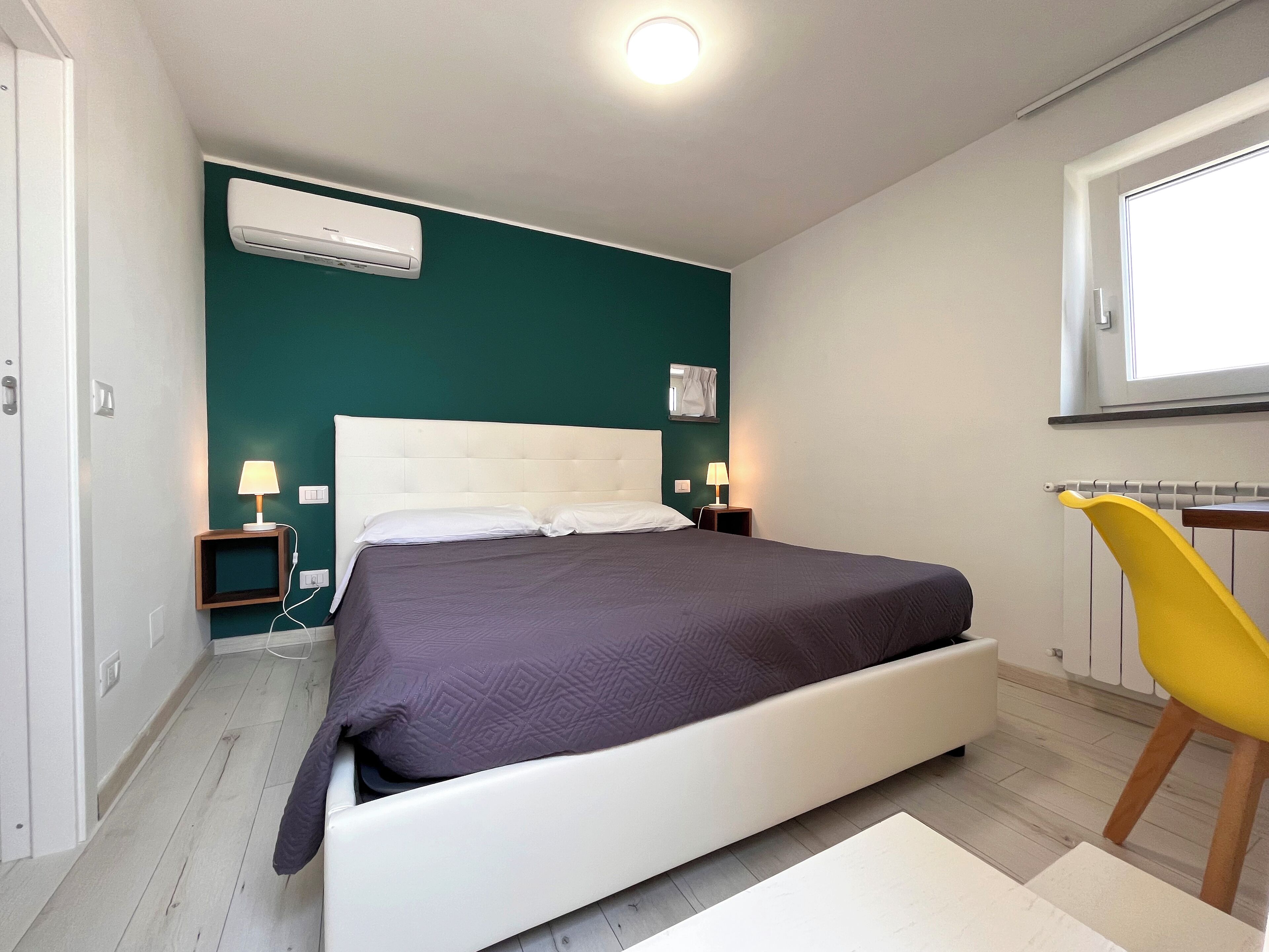 A bedroom with a bed, bedside lamps, an air conditioner, and a window.