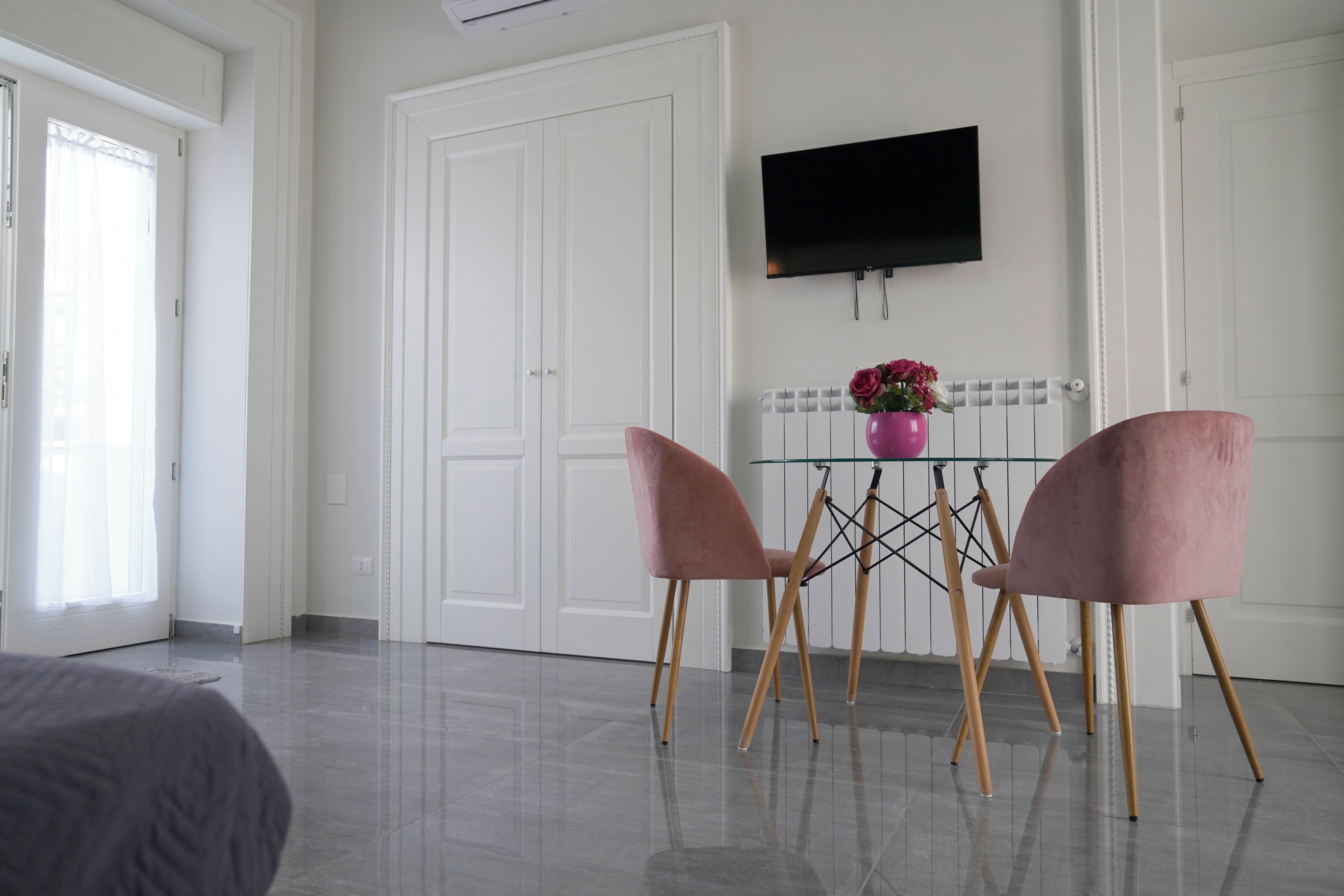 A modern room with a bed, a TV mounted on the wall, a table with chairs, and a radiator with flowers.