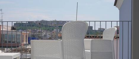 A balcony with two white wicker chairs and a small table, overlooking a cityscape.