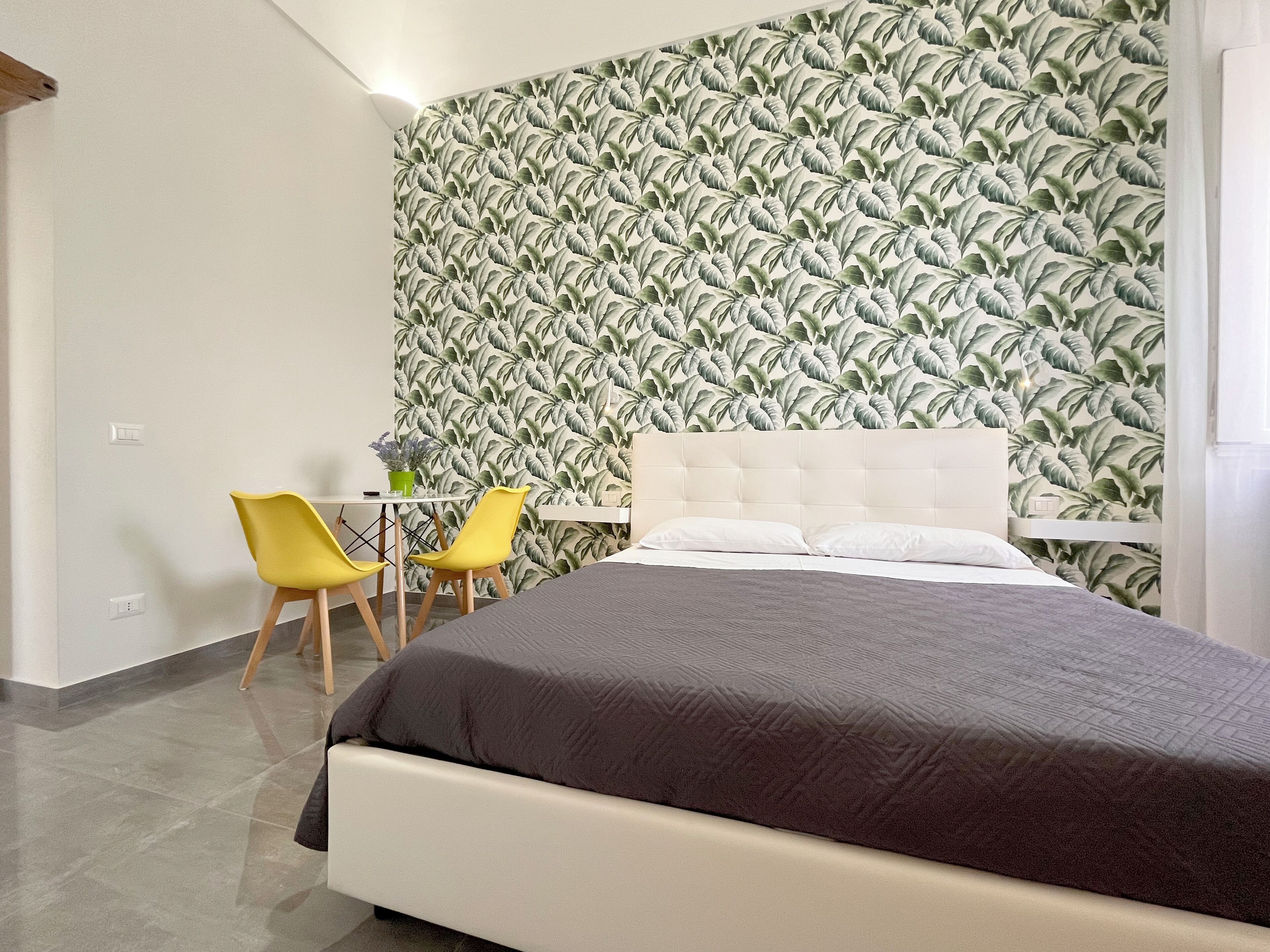 Un dormitorio con una cama, dos sillas amarillas, una mesita con una planta en maceta y una pared con un diseño de hojas.