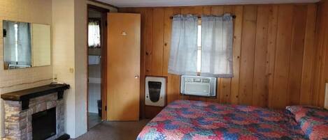 A room with a stone fireplace, a bed with a colorful quilt, wooden walls, a window with curtains, and a door leading to another room.