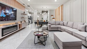 A modern living room with a large flat-screen TV, a sectional sofa, a coffee table, and a dining area in the background. - 7BR house sleeps 24 with game room (Clermont)