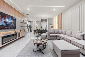 A modern living room with a large flat-screen TV, a sectional sofa, a coffee table, and a dining area in the background.