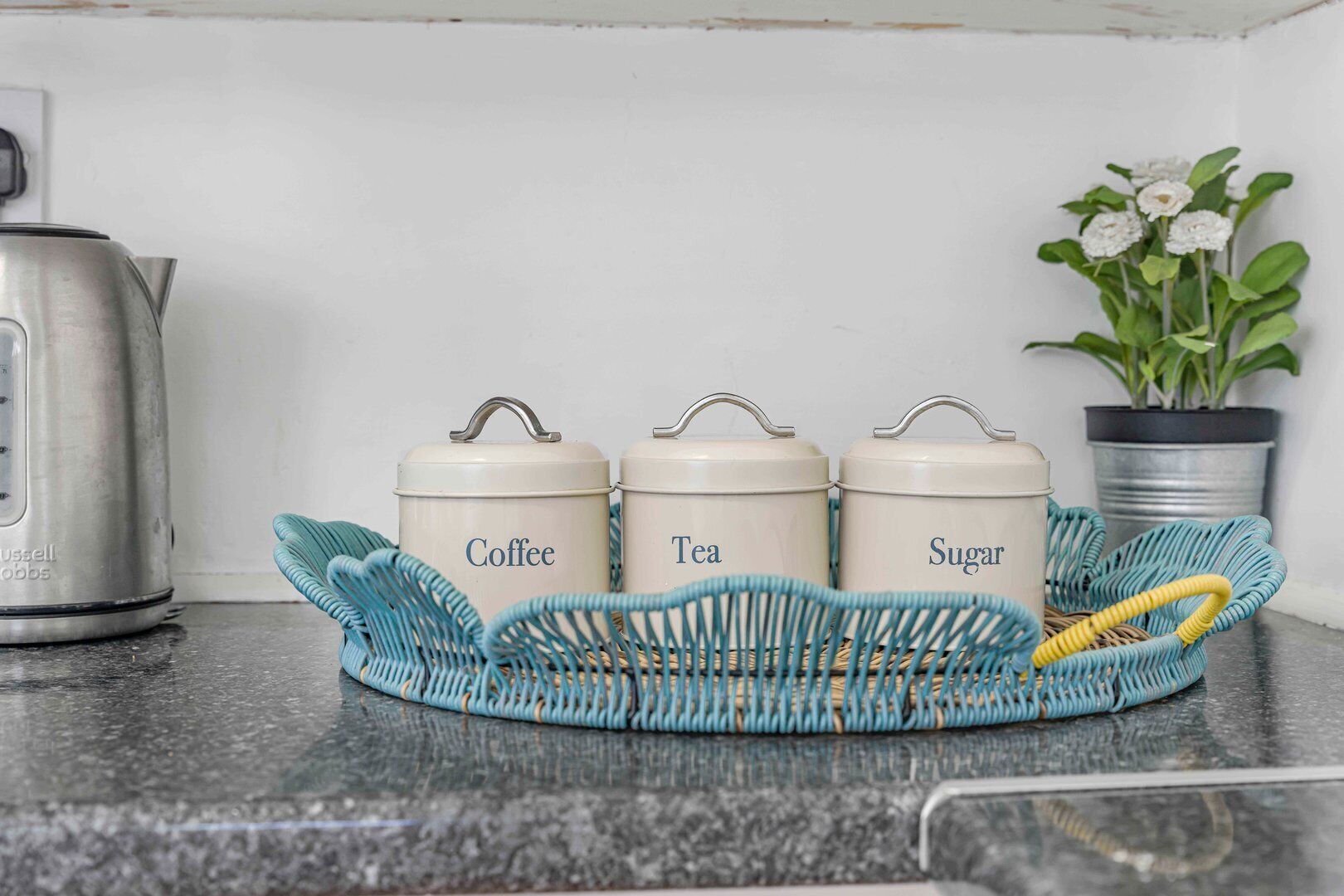 Drei beschriftete Behälter auf einer Küchenarbeitsplatte, ein Wasserkocher und ein Blumentopf.
