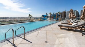 A rooftop pool area with lounge chairs and umbrellas, a clear sky, and a cityscape in the background.