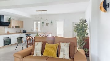 A modern living room with a brown sofa, a dining area with a table and chairs, a kitchen with white cabinets, and a large potted plant.