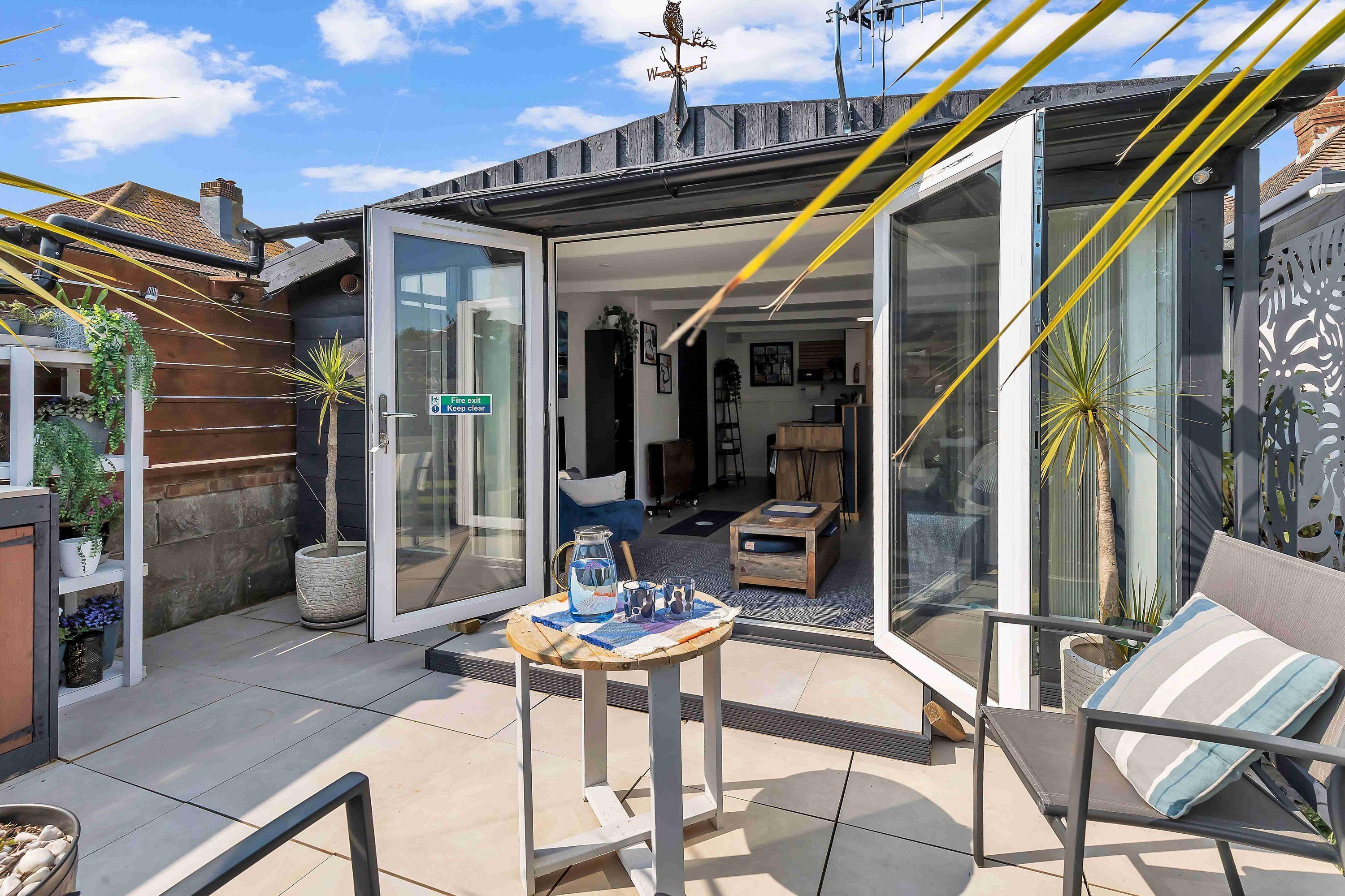 Un patio moderno con un ventanal de vidrio que da a un espacio interior, una mesa pequeña con bebidas y una silla con cojín a rayas.
