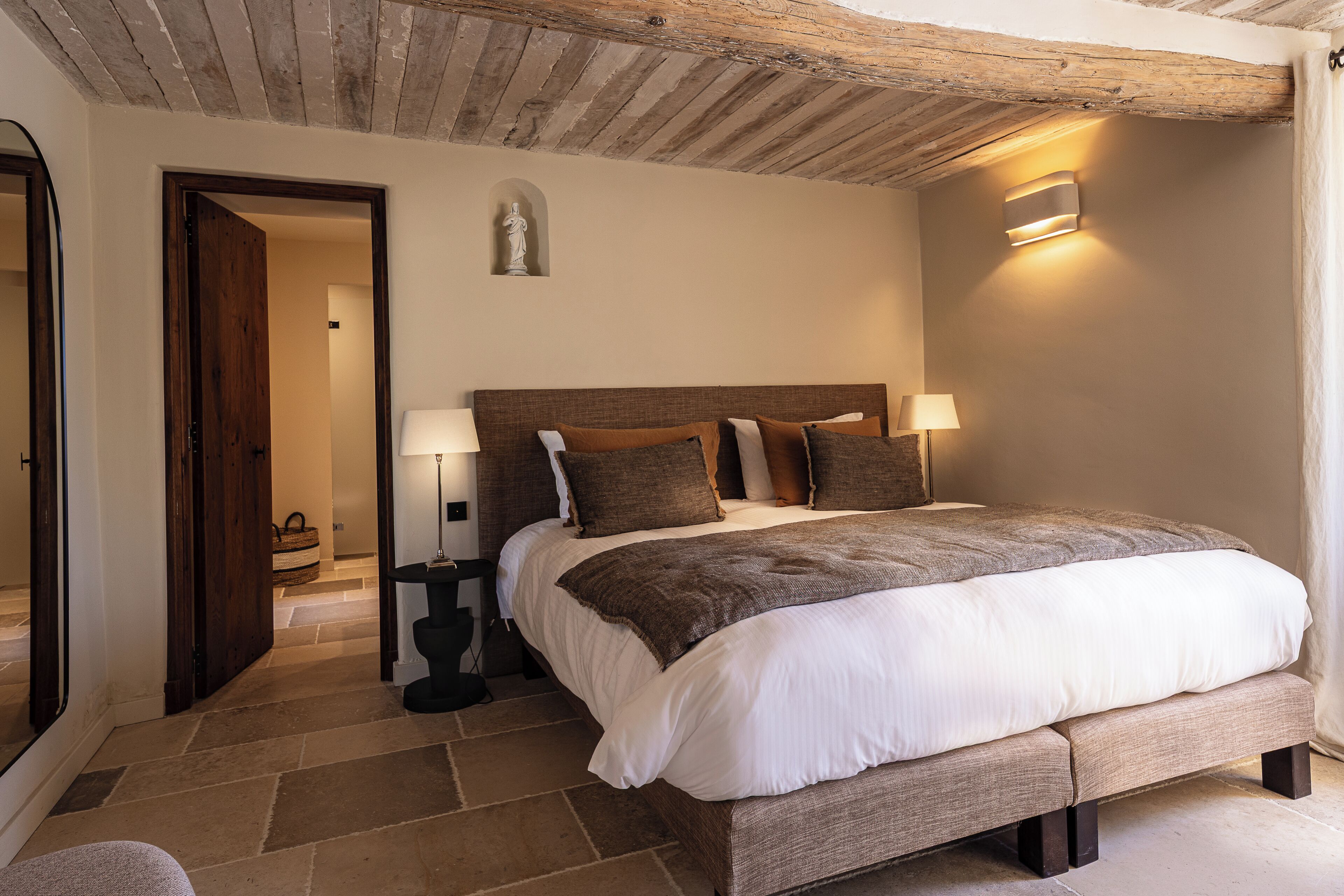 Een slaapkamer met een groot bed, twee nachtkastjes met lampen, een houten plafond en een stenen vloer.