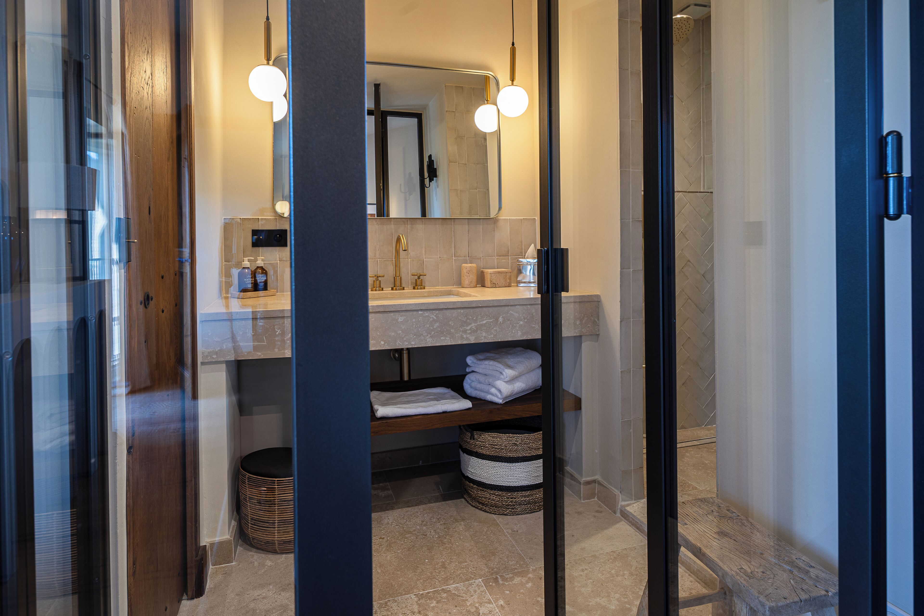 A modern bathroom with a glass shower enclosure, a wooden bench, and a vanity with a mirror and two pendant lights.