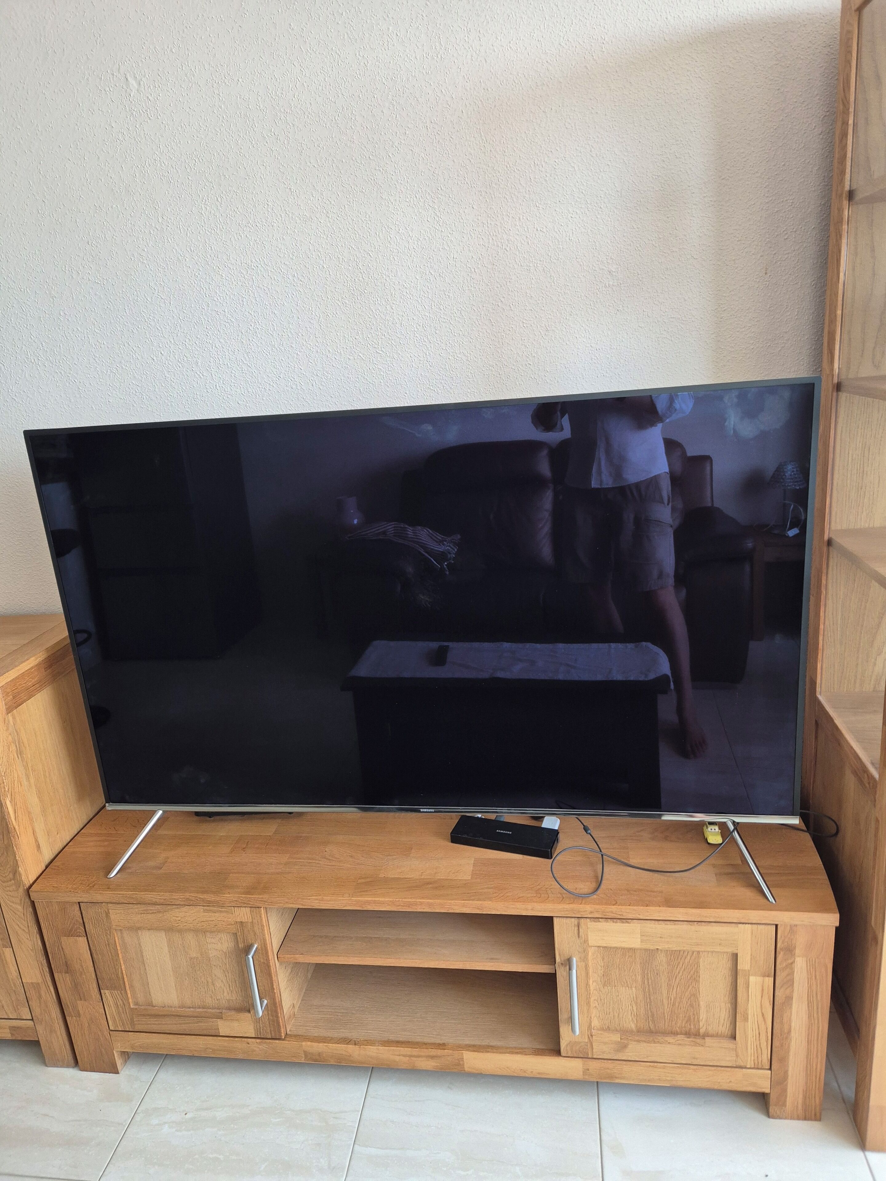 Un grand téléviseur à écran plat posé sur un meuble télé en bois, avec une télécommande et une multiprise.