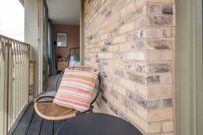 A balcony with wicker chairs and a striped cushion, a brick wall, and a glimpse of a room with a bed and a window.